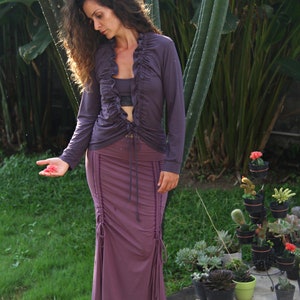 May include: A woman wearing a purple long-sleeved blouse with a ruffled neckline and a matching purple skirt. The skirt has a drawstring detail and is fitted at the waist. She is standing in front of a green leafy background.