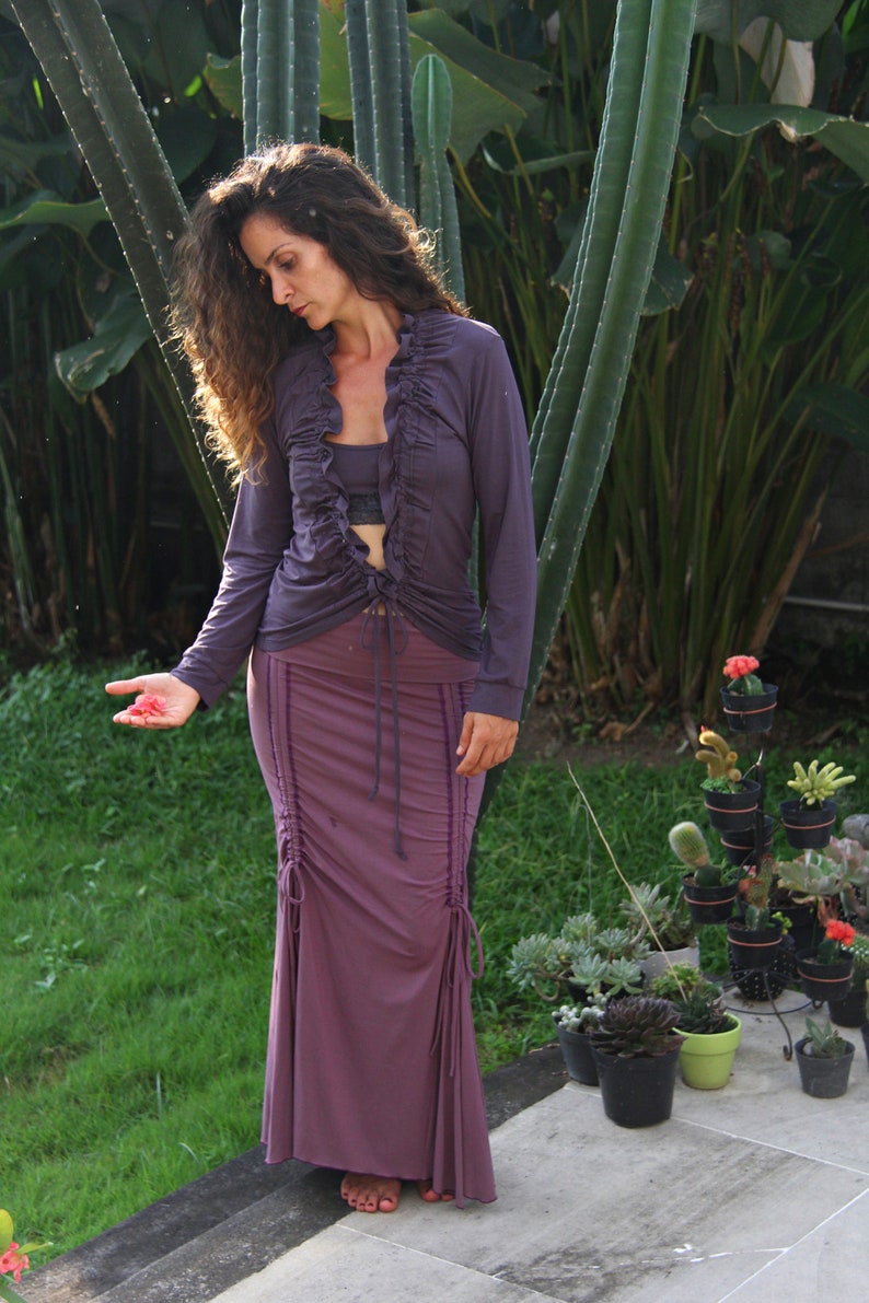 May include: A woman wearing a purple long-sleeved blouse with a ruffled neckline and a matching purple skirt. The skirt has a drawstring detail and is fitted at the waist. She is standing in front of a green leafy background.