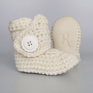 Puede incluir: Un par de botines de beb&eacute; de crochet de color crema con un bot&oacute;n blanco en la parte superior de un bot&iacute;n. El otro bot&iacute;n tiene una suela de cuero crema con la letra "R" grabada.