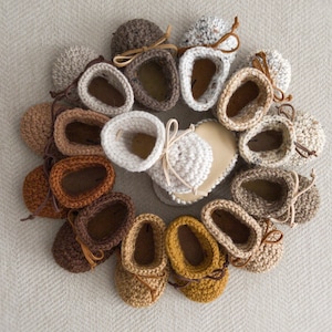 May include: A collection of 13 crocheted baby booties in various shades of brown, beige, and cream. The booties have a simple design with a rounded toe and a small bow at the top.