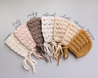 May include: Seven crocheted baby bonnets in various neutral colors, including cream, dusty pink, rocky road, oatmeal, wheat, almond, and ginger. Each bonnet has a tie at the back.