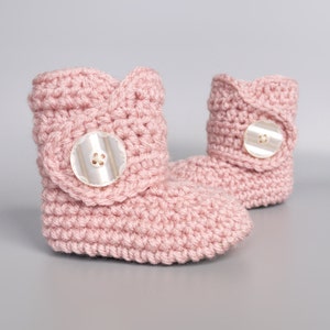 May include: A pair of pink crocheted baby booties with a button on each. The booties are made with a soft, textured yarn.