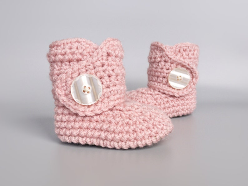 May include: A pair of pink crocheted baby booties with a button on each. The booties are made with a soft, textured yarn.