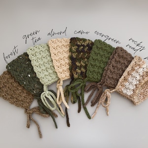 May include: A set of ten crocheted baby bonnets in various shades of green, brown, and beige. The bonnets are arranged in a semi-circle with the names of each color written above them. The colors are: bark, forest, green tea, almond, camo, evergreen, rocky road, and pebble.