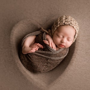 May include: A newborn baby is swaddled in a brown knitted blanket and wearing a crocheted hat. The baby is sleeping peacefully on a brown background.