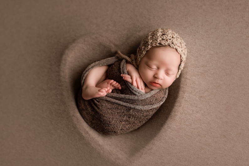 May include: A newborn baby is swaddled in a brown knitted blanket and wearing a crocheted hat. The baby is sleeping peacefully on a brown background.