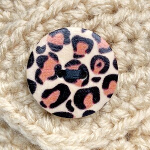 May include: A close-up of a beige crocheted fabric with a round, wooden button featuring a pink and black leopard print.