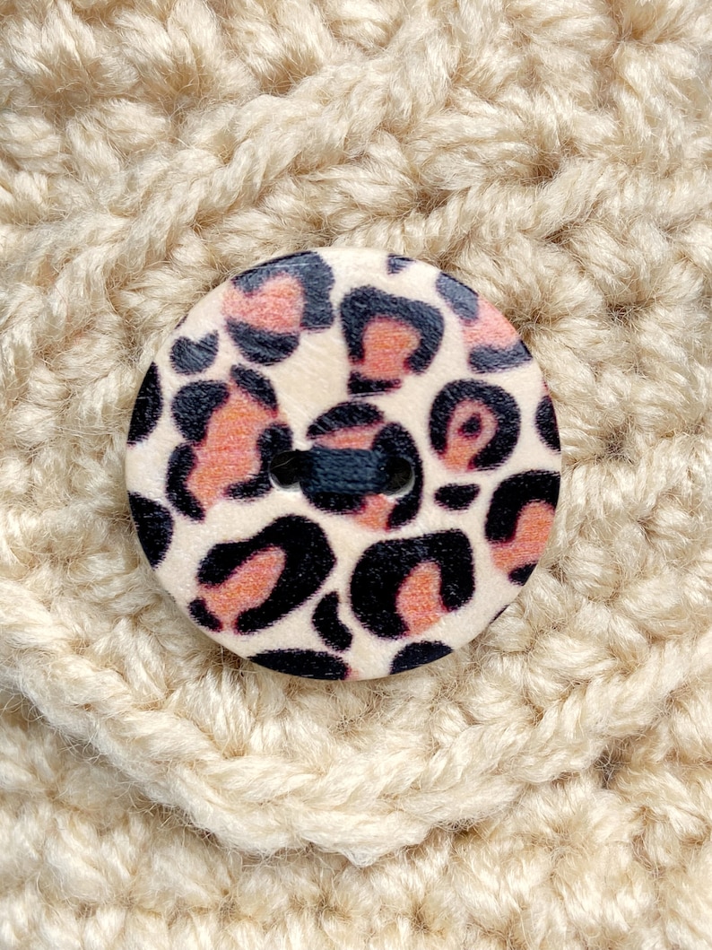 May include: A close-up of a beige crocheted fabric with a round, wooden button featuring a pink and black leopard print.