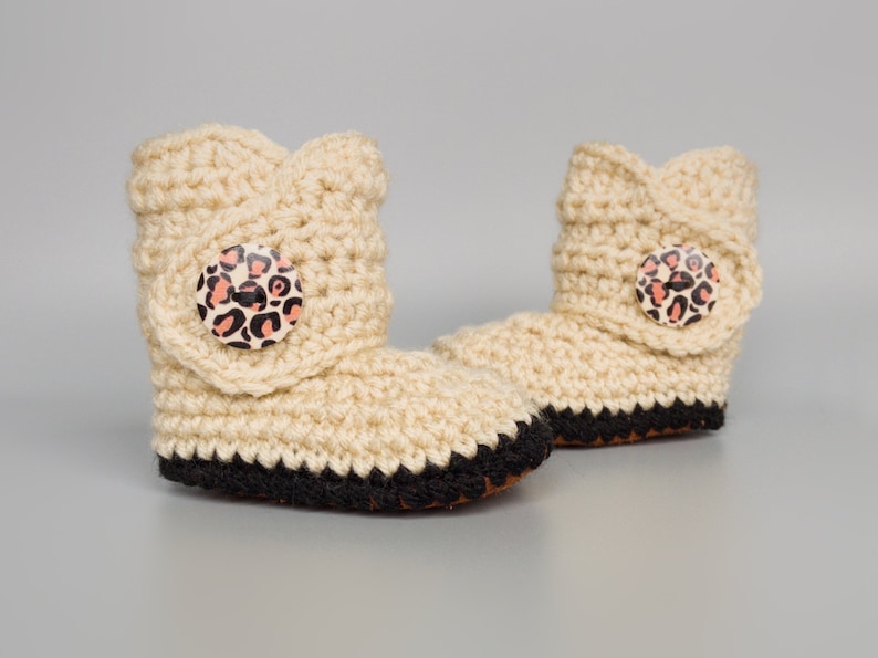May include: A pair of beige crocheted baby booties with black trim and leopard print buttons.