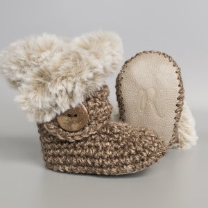 May include: A pair of brown crocheted baby booties with faux fur trim and a wooden button. The booties have a leather sole with the letter "R" embossed on it.