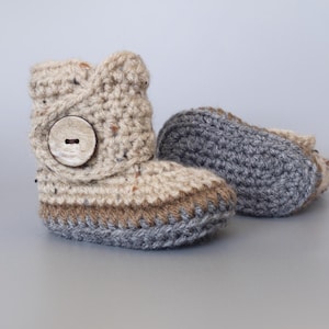 May include: A pair of crocheted baby booties with a brown button. One bootie is beige with brown and gray trim, the other is gray.