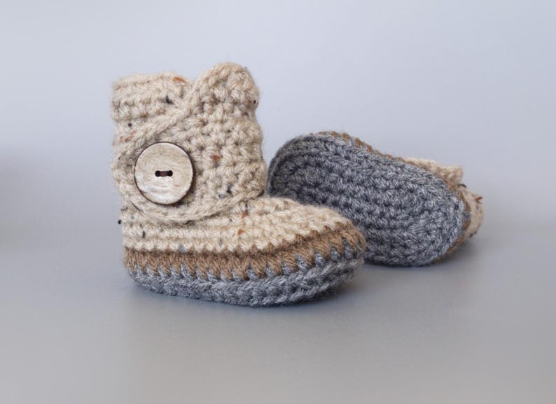 May include: A pair of crocheted baby booties with a brown button. One bootie is beige with brown and gray trim, the other is gray.