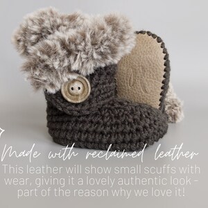 May include: A dark gray crochet baby bootie with a faux fur trim and a button closure. The bootie has a light brown leather sole. The text "Made with reclaimed leather" is visible in the image.