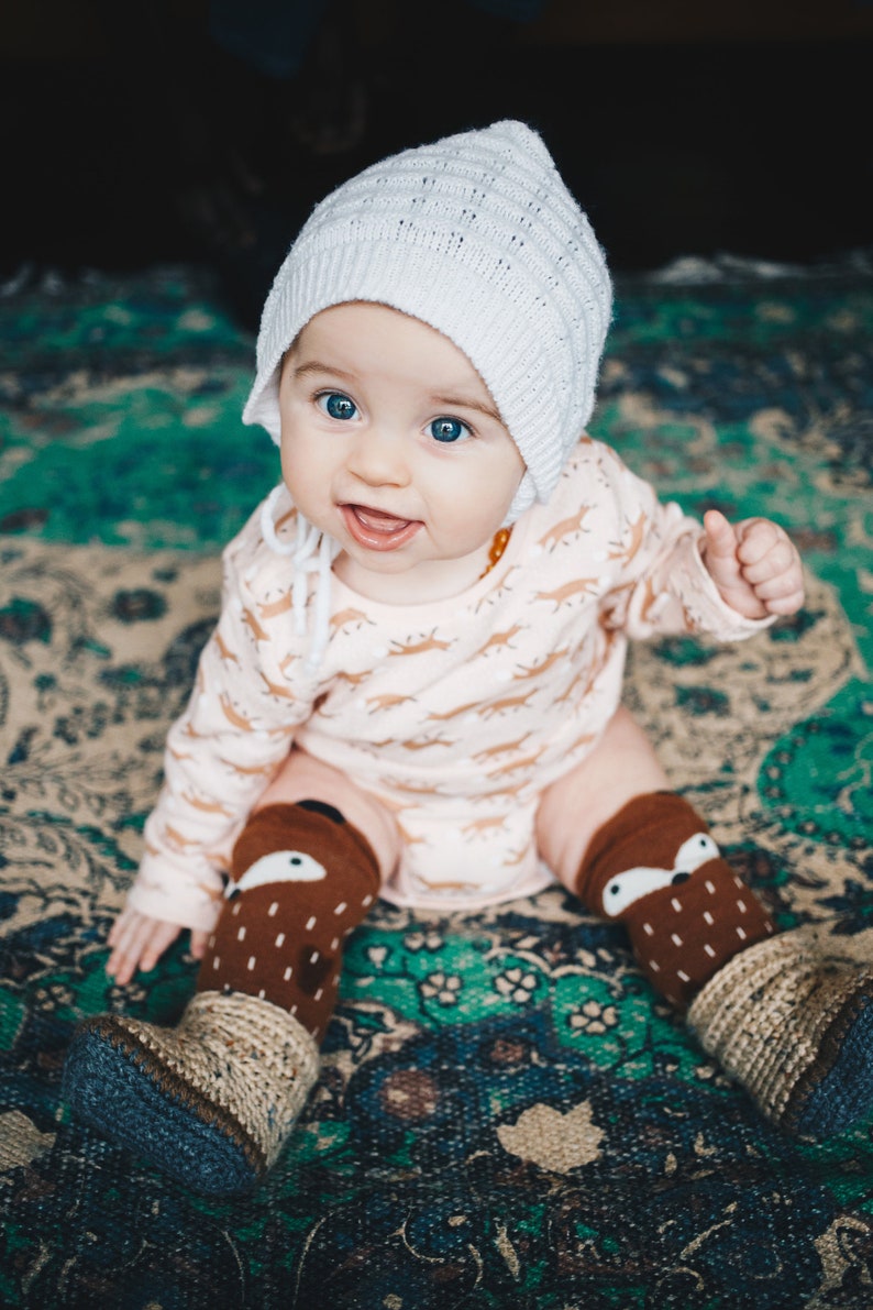 May include: A baby wearing a white knitted bonnet, a pink long-sleeved shirt with a brown animal print, and brown and white knitted leg warmers with a fox design. The baby is sitting on a colorful rug.