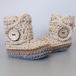 May include: A pair of beige crocheted baby booties with brown and gray trim. Each bootie has a large wooden button on the side.