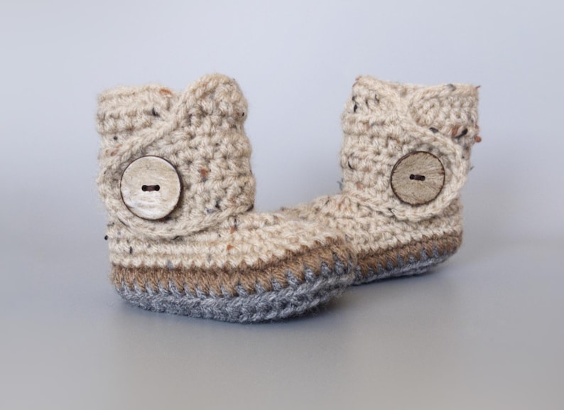 May include: A pair of beige crocheted baby booties with brown and gray trim. Each bootie has a large wooden button on the side.