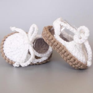 May include: A pair of white crocheted baby sandals with brown jute soles. The sandals have a simple design with a single strap across the top of the foot.