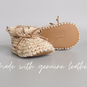 May include: A pair of beige crocheted baby booties with brown leather laces and soles. The booties are made with genuine leather and have a soft, comfortable feel.