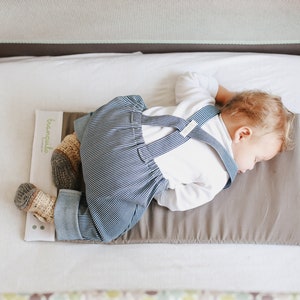 May include: A baby sleeping on a gray and white portable crib mattress. The baby is wearing a white shirt and blue striped overalls. The mattress has a brown waterproof liner with the word "breathable" printed on it.