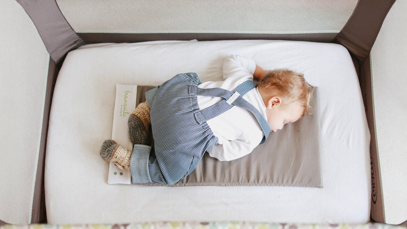 May include: A baby sleeping on a gray and white portable crib mattress. The baby is wearing a white shirt and blue striped overalls. The mattress has a brown waterproof liner with the word "breathable" printed on it.
