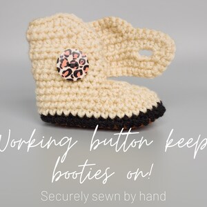 May include: Cream crocheted baby booties with a black trim and a leopard print button. The text "Working button keeps booties on! Securely sewn by hand" is below the booties.