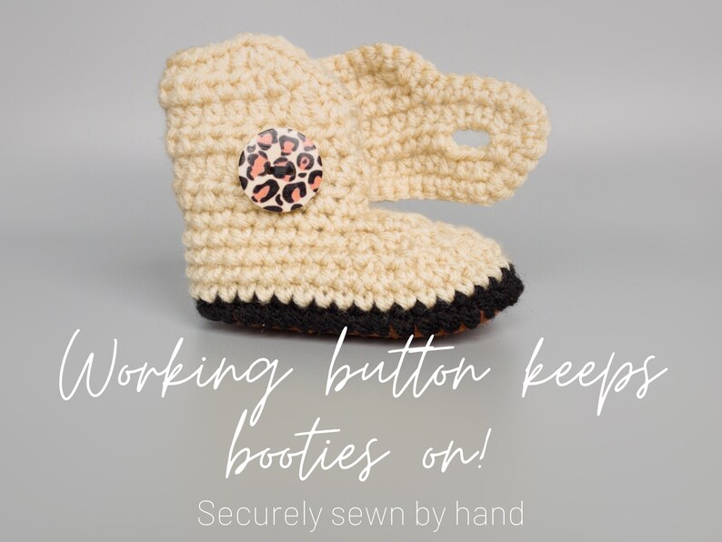 May include: Cream crocheted baby booties with a black trim and a leopard print button. The text "Working button keeps booties on! Securely sewn by hand" is below the booties.