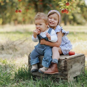 May include: Two children, a girl and a boy, sit on a wooden crate in a field. The girl is wearing a light purple dress with white polka dots and a pink and white headband. The boy is wearing a blue and white striped overall and a white shirt. They are both smiling and looking at the camera.