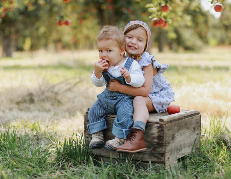 May include: Two children, a girl and a boy, sit on a wooden crate in a field. The girl is wearing a light purple dress with white polka dots and a pink and white headband. The boy is wearing a blue and white striped overall and a white shirt. They are both smiling and looking at the camera.