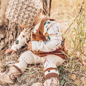 May include: A baby wearing a brown crocheted hat, a white long-sleeved shirt with a woodland print, a brown corduroy vest, brown and white leggings, and brown boots. The baby is sitting in tall grass.
