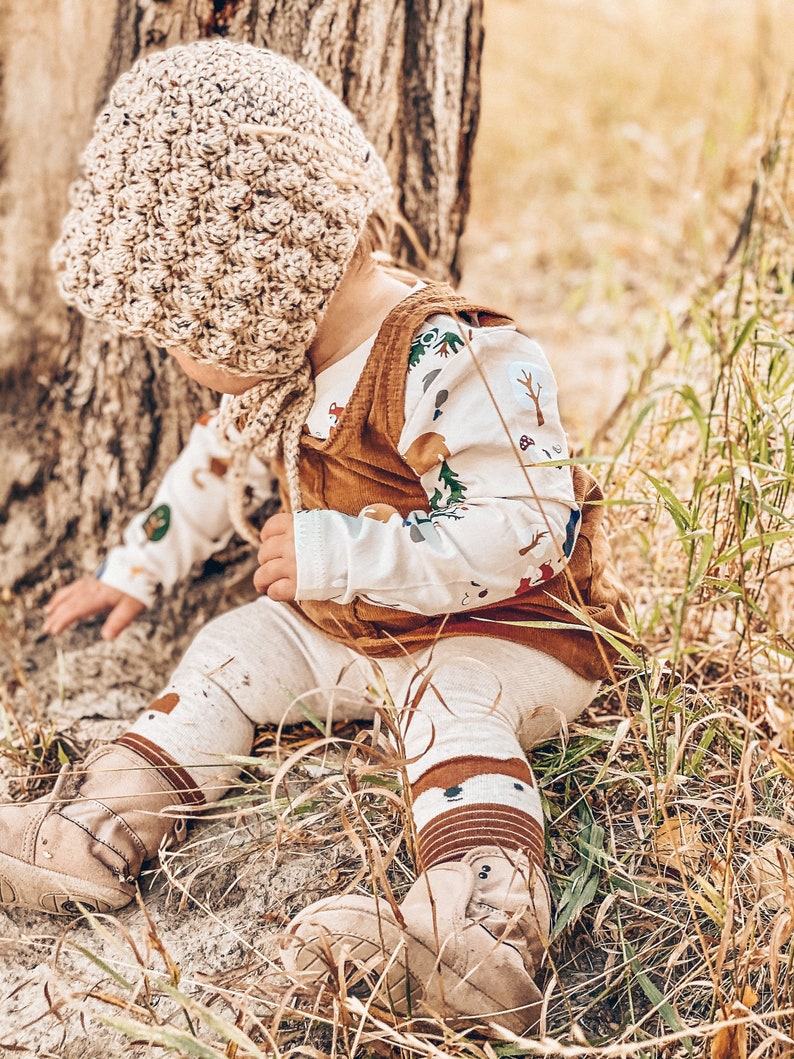 May include: A baby wearing a brown crocheted hat, a white long-sleeved shirt with a woodland print, a brown corduroy vest, brown and white leggings, and brown boots. The baby is sitting in tall grass.