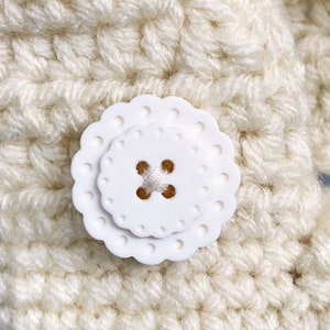 Puede incluir: Una tela de crochet blanca con un bot&oacute;n de pl&aacute;stico blanco con borde festoneado.