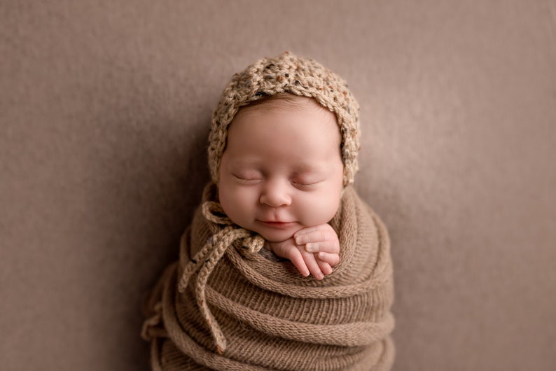 May include: A newborn baby is swaddled in a brown knitted blanket and wearing a crocheted hat. The baby is sleeping peacefully with their hands tucked in.