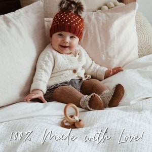 May include: A baby wearing a white cardigan, brown tights, and a red knitted hat with a white pom-pom sits on a white bed with a wooden ring toy. The text "100% Made with Love!" is written on the bed.