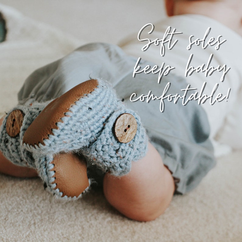 May include: A baby wearing soft blue knitted booties with brown leather soles and wooden buttons. The booties say "Soft soles keep baby comfortable!"