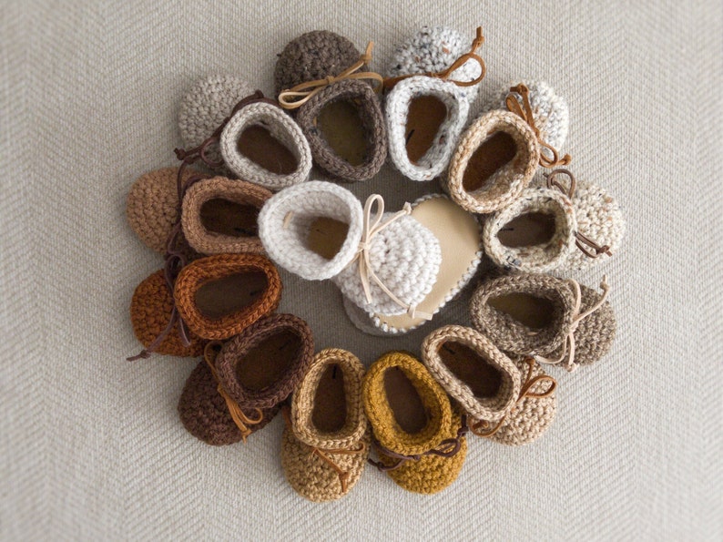 May include: A collection of 13 crocheted baby booties in various shades of brown, beige, and cream. The booties have a simple design with a rounded toe and a small bow at the top.