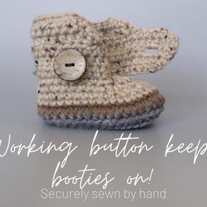 May include: Crocheted baby booties with a brown button and a gray trim. The text "Working button keeps booties on! Securely sewn by hand" is below the booties.