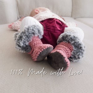 May include: A pair of pink crocheted baby booties with gray faux fur trim and brown suede soles. The booties are on a white couch with a baby lying on it. The text "100% Made with Love!" is written on the couch.