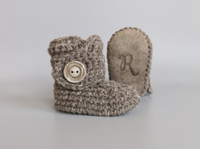 May include: A pair of brown crocheted baby booties with a button on the front and a brown suede bootie with the letter "R" embroidered on it.
