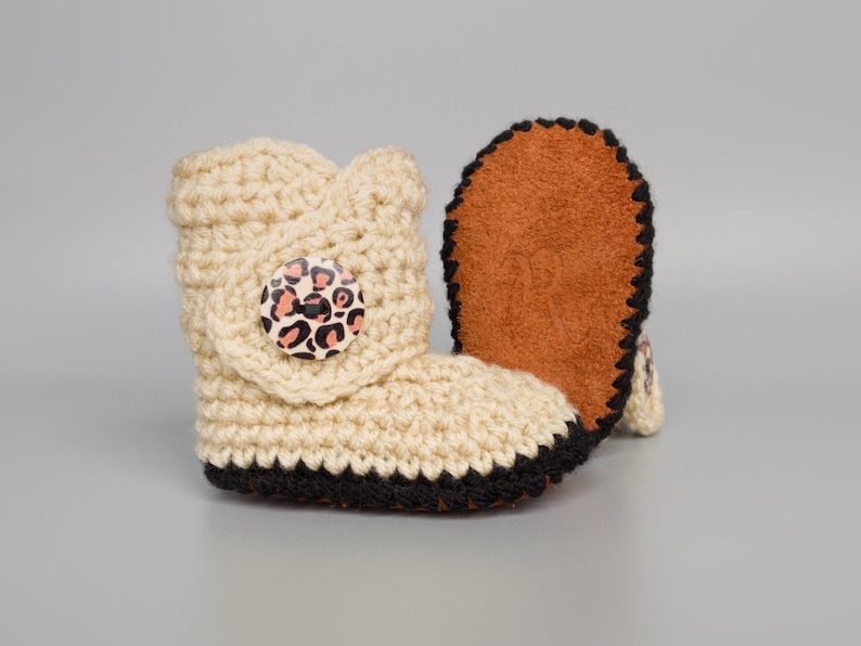 May include: A pair of beige crocheted baby booties with black trim and a leopard print button. The booties have a brown suede sole.