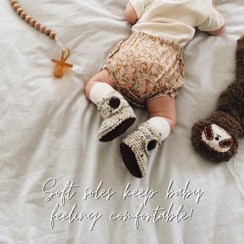 May include: A baby wearing a floral print romper and crocheted booties with soft soles. The baby is lying on a white sheet with a wooden bead necklace and a pacifier nearby. The text "Soft soles keep baby feeling comfortable!" is written on the bottom of the image.
