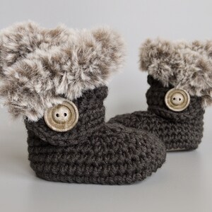 May include: A pair of dark gray crochet baby booties with faux fur trim. Each bootie features a large, round, light-colored button. The booties are designed to keep a baby's feet warm and cozy.
