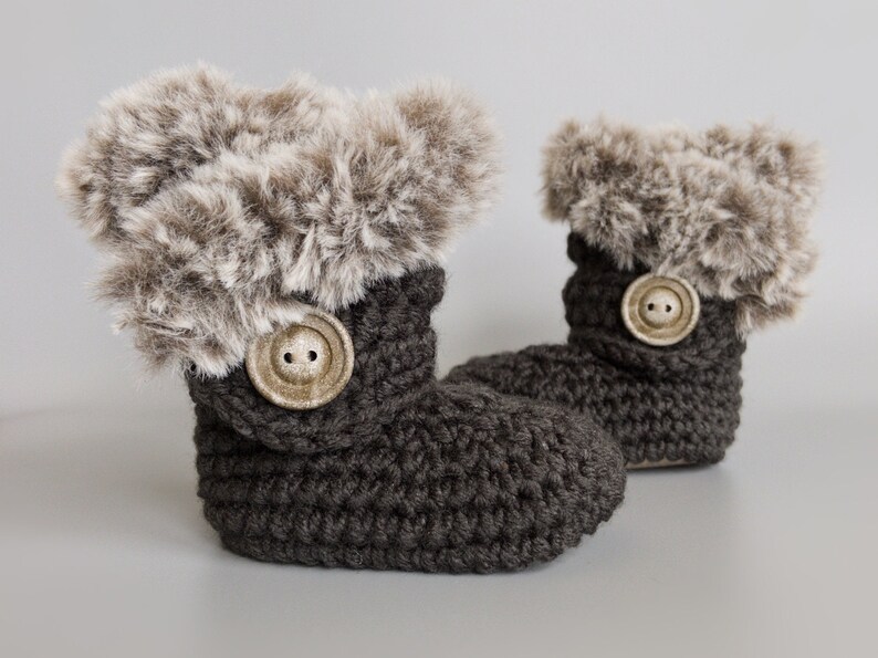 May include: A pair of dark gray crochet baby booties with faux fur trim. Each bootie features a large, round, light-colored button. The booties are designed to keep a baby's feet warm and cozy.