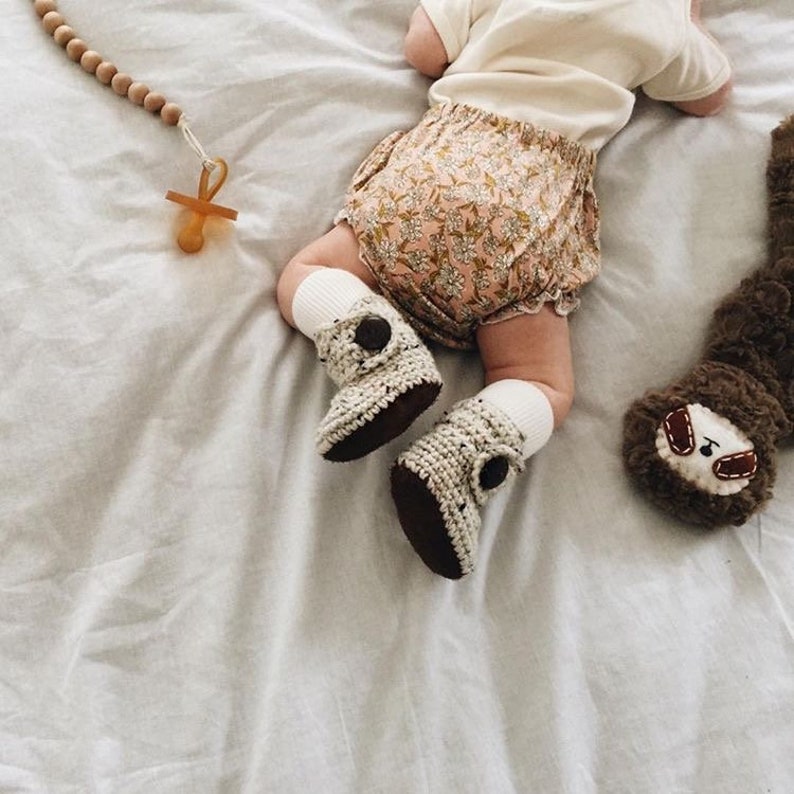 Puede incluir: Un beb&eacute; con un pa&ntilde;al de tela con estampado floral y botines de crochet con botones marrones. El beb&eacute; est&aacute; acostado en una s&aacute;bana blanca con un collar de cuentas de madera y un chupete.