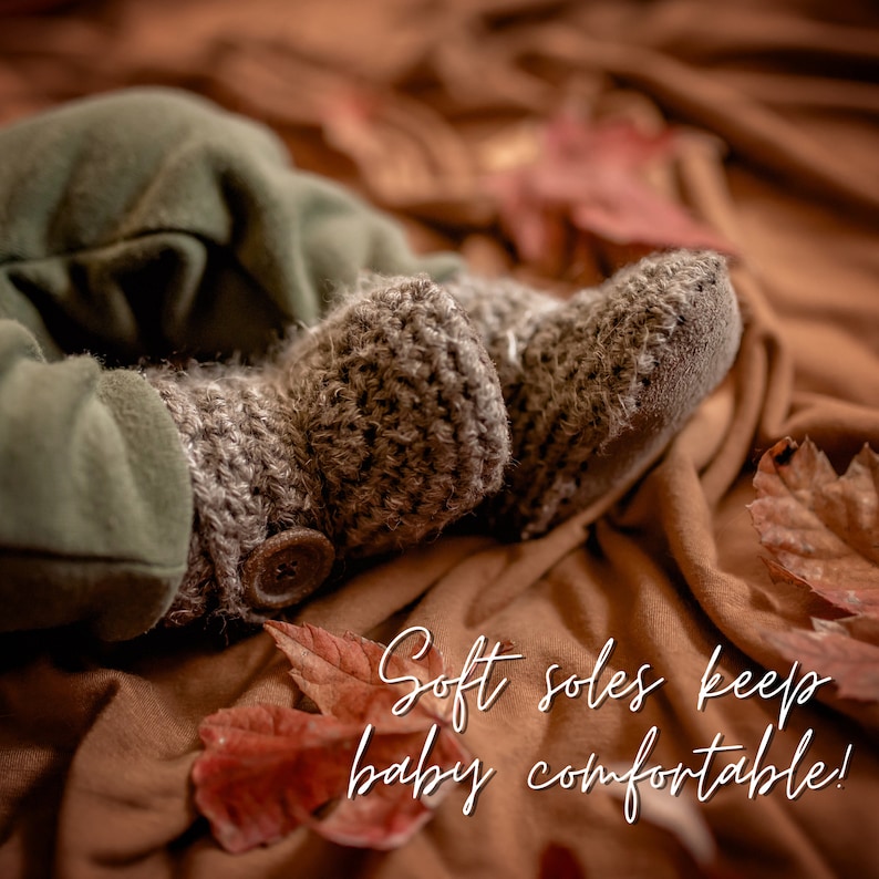 May include: Close-up of a baby's feet wearing soft, brown crocheted booties with a button detail. The booties are on a brown fabric background with fall leaves. The text "Soft soles keep baby comfortable!" is written in white cursive.