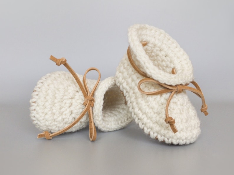 May include: A pair of white crocheted baby booties with brown leather ties. The booties have a simple design and are tied with a bow.