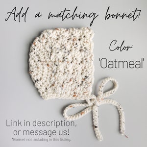 May include: A crocheted bonnet in a cream color with brown and gray flecks. The bonnet has a tie at the bottom and the text 'Color: 'Oatmeal'' is visible in the image.