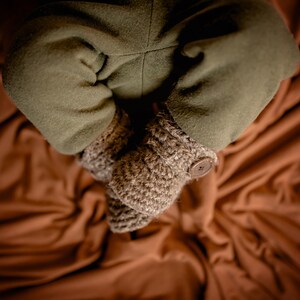 May include: A pair of brown crocheted baby booties with a button on one bootie. The booties are on a brown fabric background.
