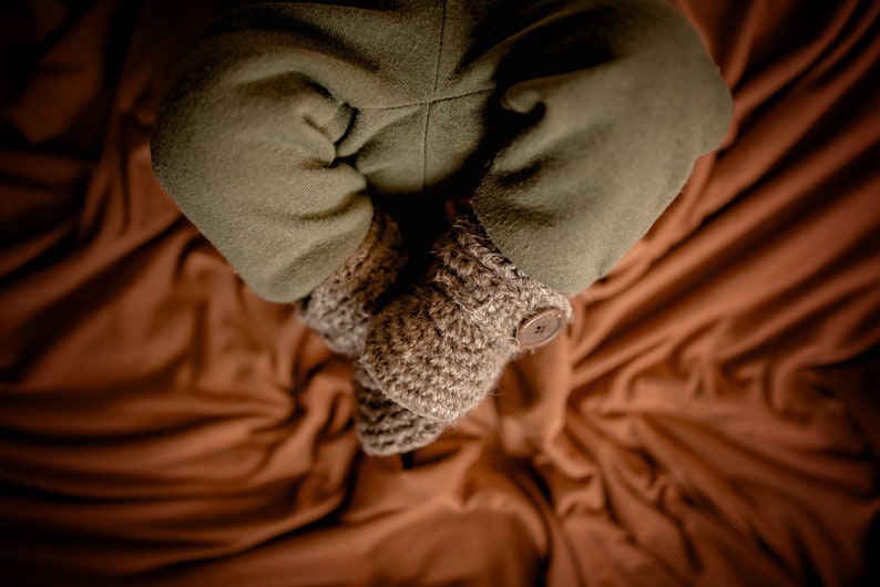 May include: A pair of brown crocheted baby booties with a button on one bootie. The booties are on a brown fabric background.