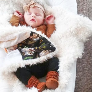 以下が含まれることがあります： 白いふわふわの毛布の上で眠っている赤ちゃん。赤ちゃんは、茶色の長袖ロンパース、黒いズボン、茶色のベビーシューズを履いています。茶色のリボン付きヘッドバンドをしています。赤ちゃんは、黒と白の柄の毛布に包まれています。毛布には花柄のデザインが施されています。