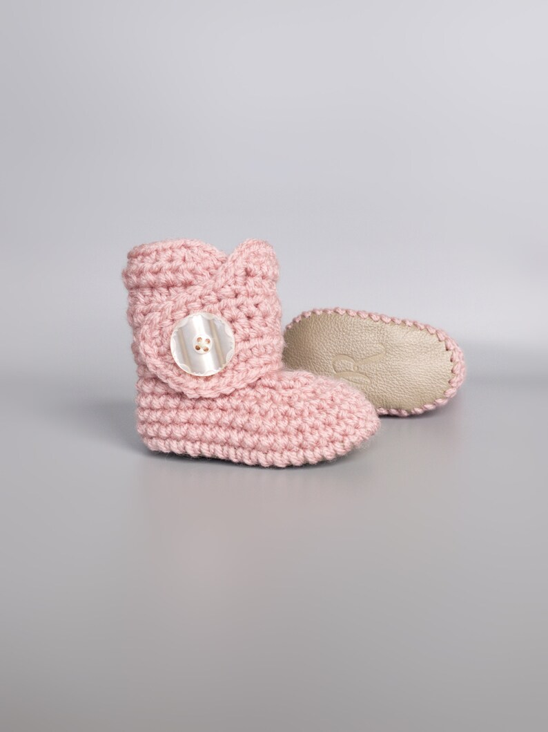 May include: A pair of pink crocheted baby booties with a white button on the top of each bootie.